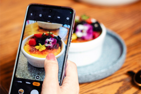 Hand holding smartphone capturing an image of a white ramekin filled with cr�me br�l�e topped with edible flowers and berries, placed on a round blue plate on a wooden table.