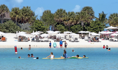 Families and children swimming and playing in the shallow water in front of a sandy beach lined with lounge chairs and umbrellas, backed by lush tropical trees.